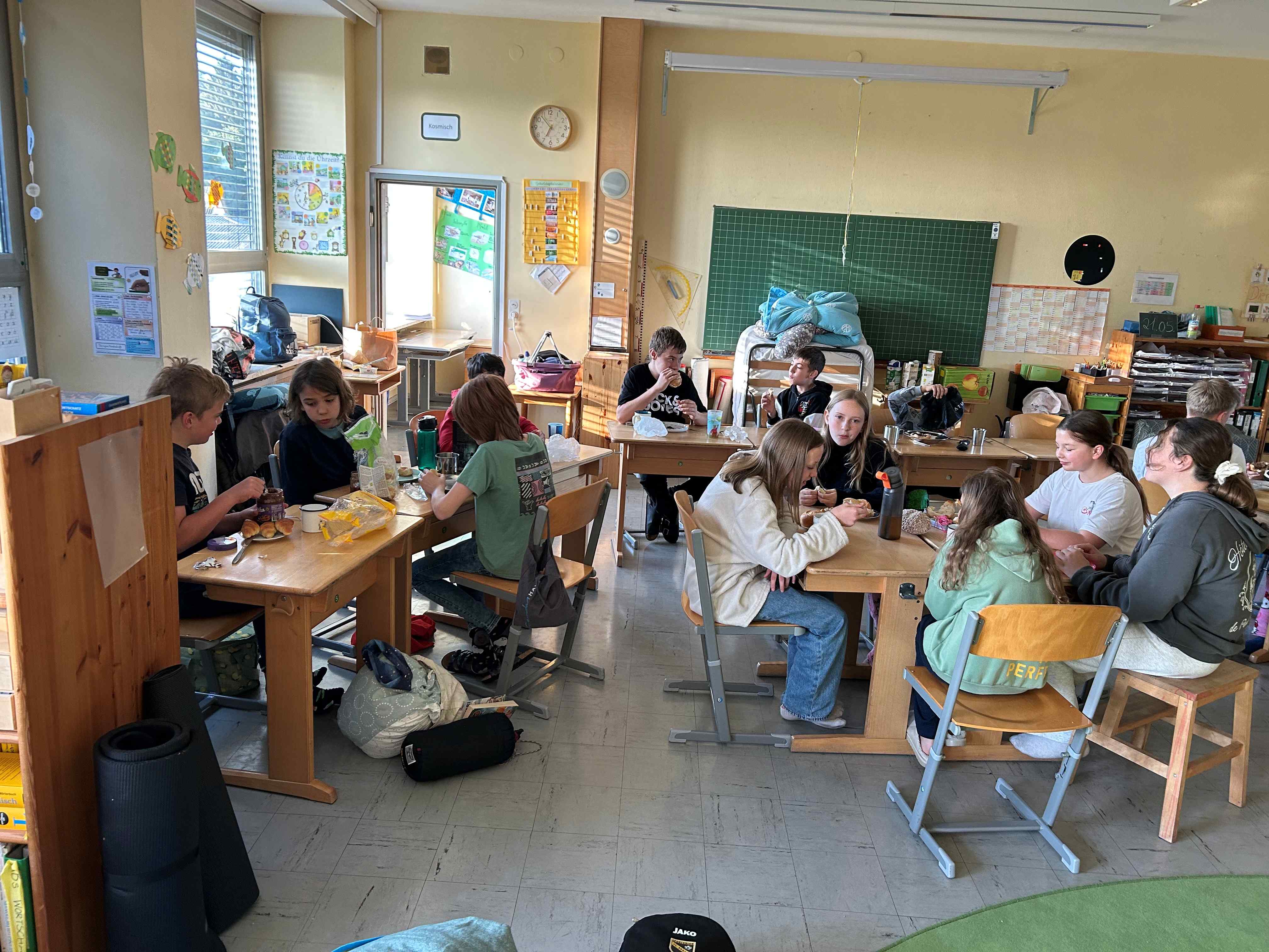 In einem Klassenzimmer sitzen Kinder verteilt an Tischen und frühstücken.