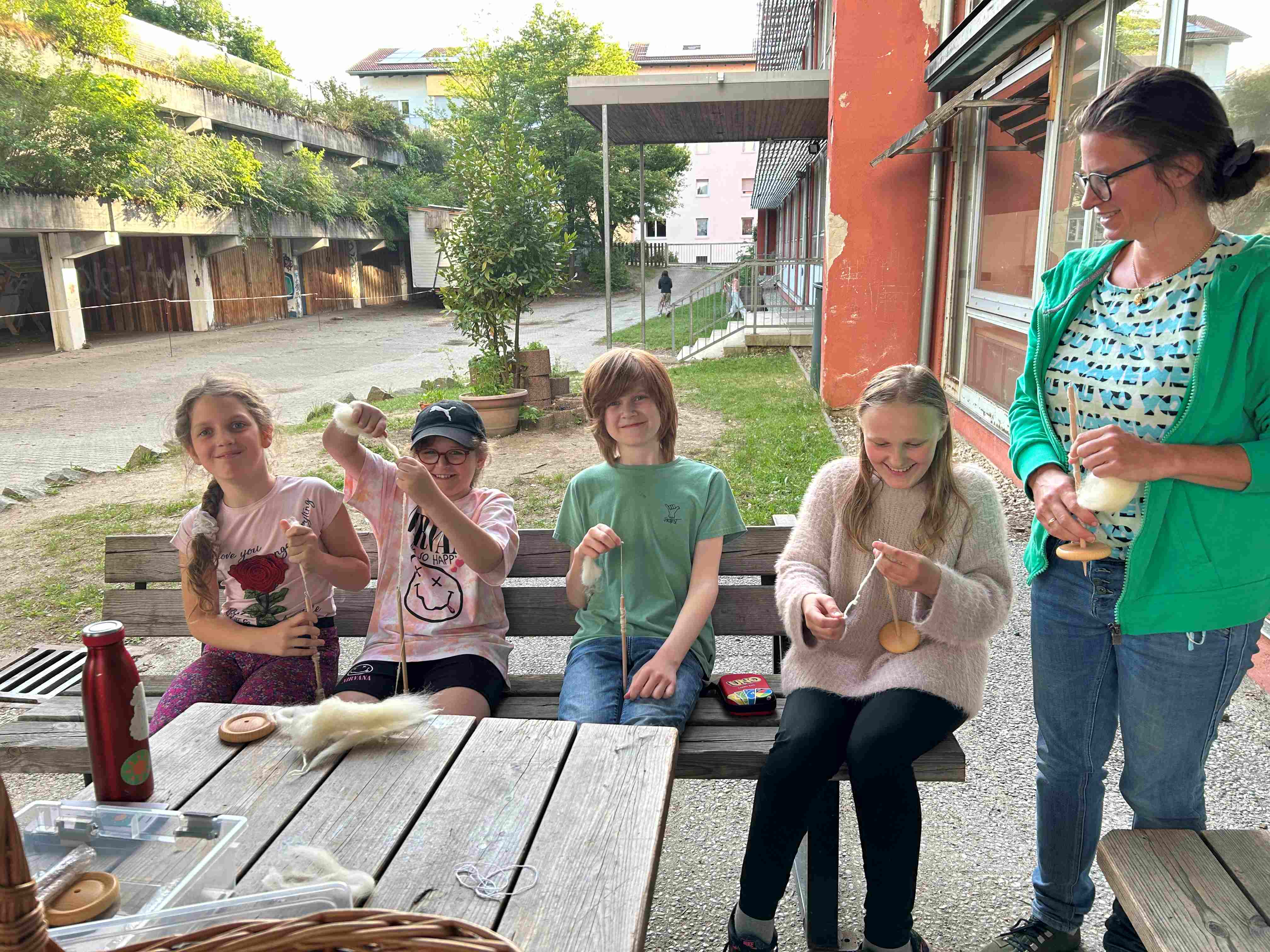 Vier Kinder sitzen auf einer Bank und spinnen mit der Hand Wolle. Daneben steht eine Frau.